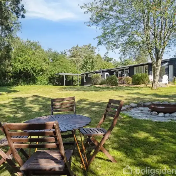 Et rundt træbord med fire træstole står på en græsplæne. I baggrunden ses et sortmalet hus og en bålplads omgivet af græs, træer og buske.