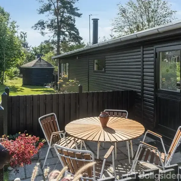 Rundt havebord med stole står på terrassen ved et sortmalet træhus. Omkring bordet er grønne planter, en lille grill, og i baggrunden ses flere træer og en grøn græsplæne.