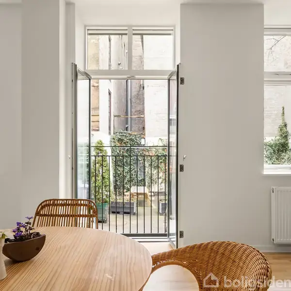 Et rundt træbord med skåle og en vase står i forgrunden. Dør åbner til en balkon med udsigt til en indre gård. Moderne indretning med store vinduer.