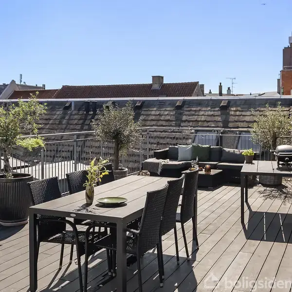 Terrasse med træbord og stole arrangeret til spisning, omgivet af plantekrukker. I baggrunden ses en sofa og et rektangulært bord på en aflang balkon med udsigt over hustage.