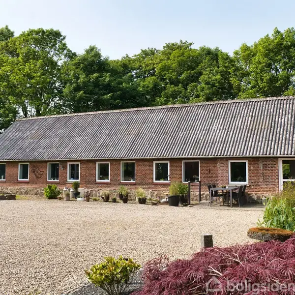 En lang, murstenslade med grått tag står i en gårdsplads omgivet af grus og planter. I baggrunden er der frodige grønne træer.