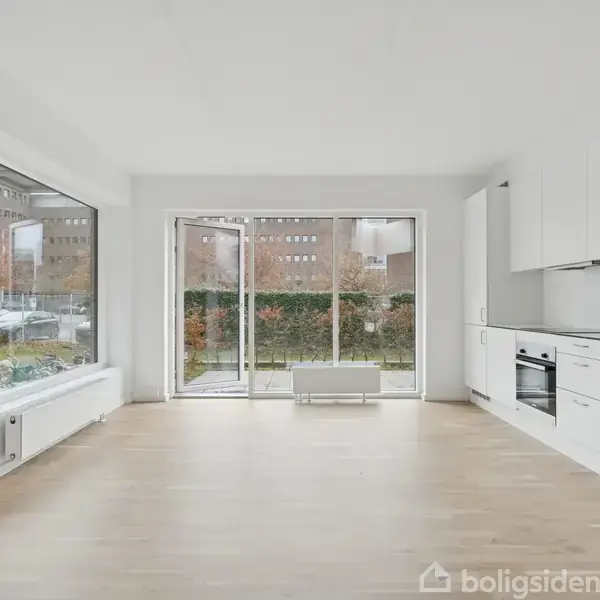 Et lyst, moderne rum med trægulv. Et hvidt køkken med indbyggede apparater er til højre. Store vinduer og en glasdør giver udsigt til en have.