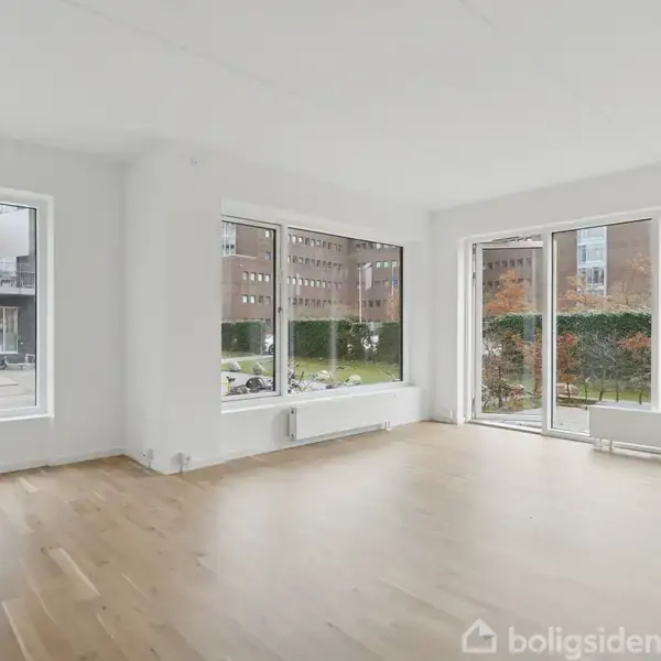 Stue med trægulv omgivet af store vinduer. Rummet er lyst og rummeligt, med udsigt til en have med buske udenfor. Ingen møbler er til stede.