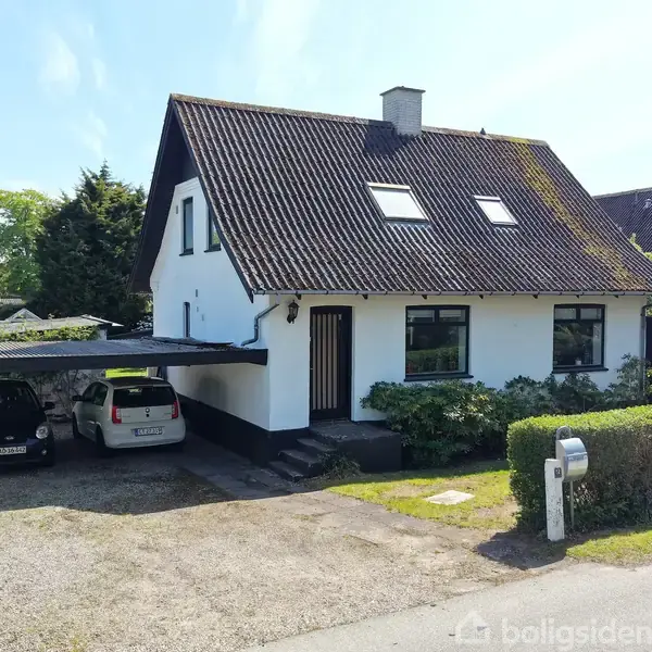 Et hvidt hus med skråt tag står i et villakvarter. To biler holder parkeret i en overdækket carport ved siden af huset. Græs, buske og en hæk omgiver ejendommen.