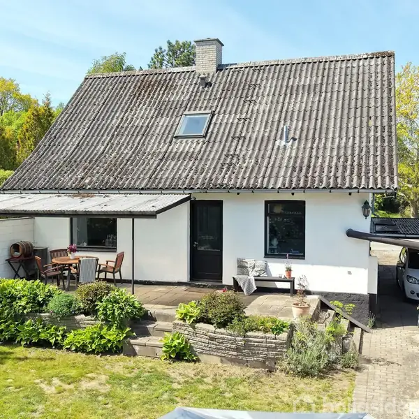 Et hvidt hus står omgivet af en have med grønne buske. En terrasse foran huset har et bord og stole. En grusvej fører til en garage på siden.