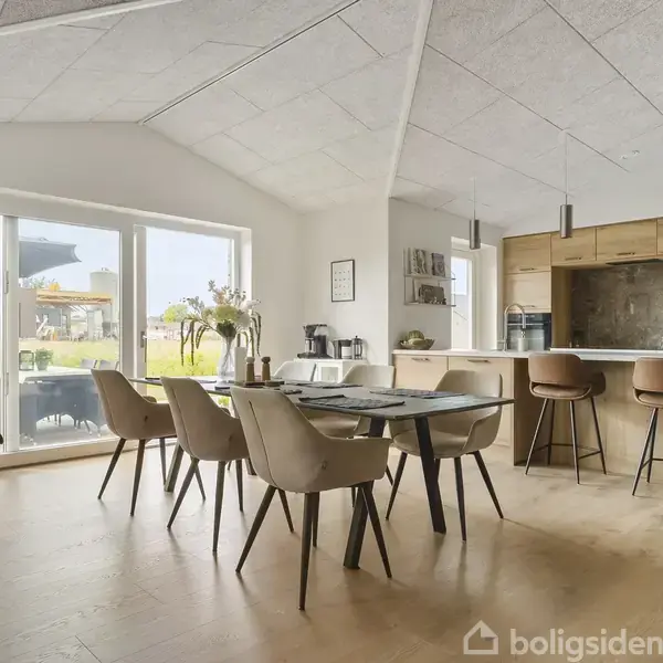 En spisebord med otte stole står i et lyst køkken-alrum med trægulv; store vinduer giver udsigt til en have. Et køkkenområde med barstole ses i baggrunden.