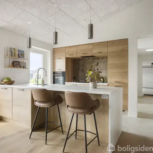 Køkken med to brune barstole ved hvid køkkenø. Træskabe, indbygget ovn, og planter i baggrunden. Moderne loftslamper hænger over øen. Lyse vægge og gulv.