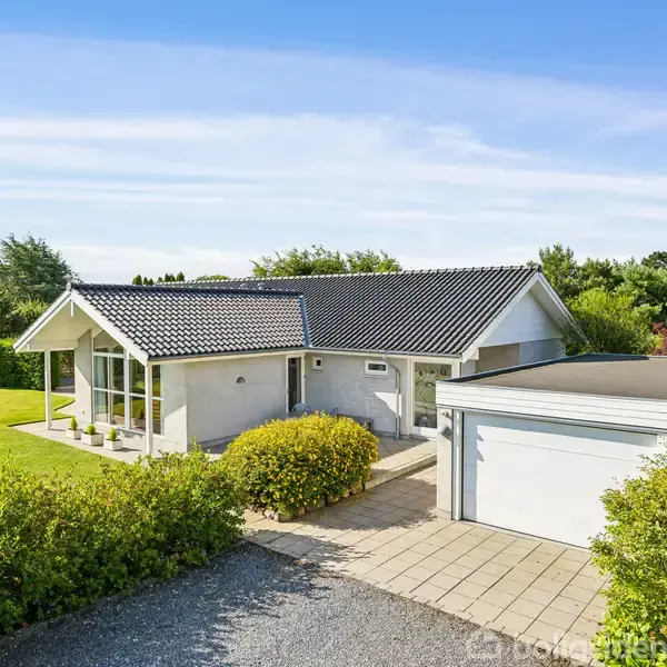 En moderne villa med sort tag ligger i en velplejet have. Ved siden af er der en hvid garage. Omkring huset er der grønne træer og buske.