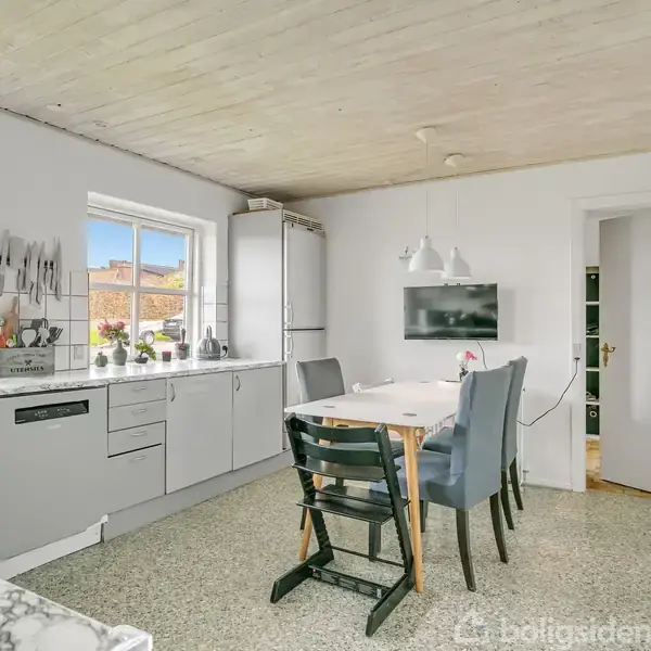 Køkken med grå skabe, marmor bordplade og spiseplads. To vinduer giver lys. En dør fører til et andet rum. Indretningen er moderne med minimalistiske lamper.