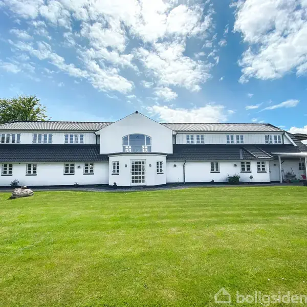 En stor, hvid to-etagers bygning står midt i en velplejet grøn græsplæne med en blå himmel og spredte skyer som baggrund.