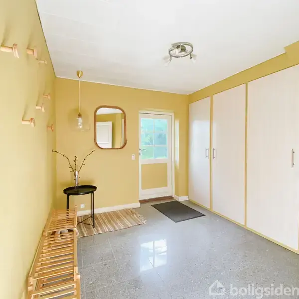 Entré med gul væg, skoopbevaring og knagerække. Lampe hænger ned fra loftet. Døren med vindue leder udenfor. Skabe på højre væg, spejl ved siden af lampen.