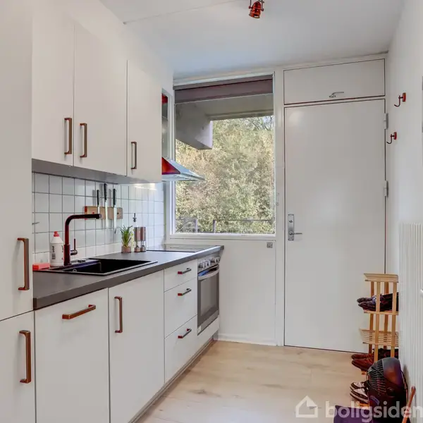 Et hvidt, moderne køkken har sorte greb og en vask, set fra siden. Et komfur står ved vinduet med udsigt til grønne trætoppe. Spiseplads til højre.