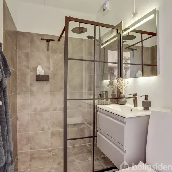 Bruseområdet adskilt af glas med sorte rammer, står ved siden af en håndvask med skuffer. Toilet i forgrunden, væggen har lyse fliser. Håndklæder hænger til venstre.