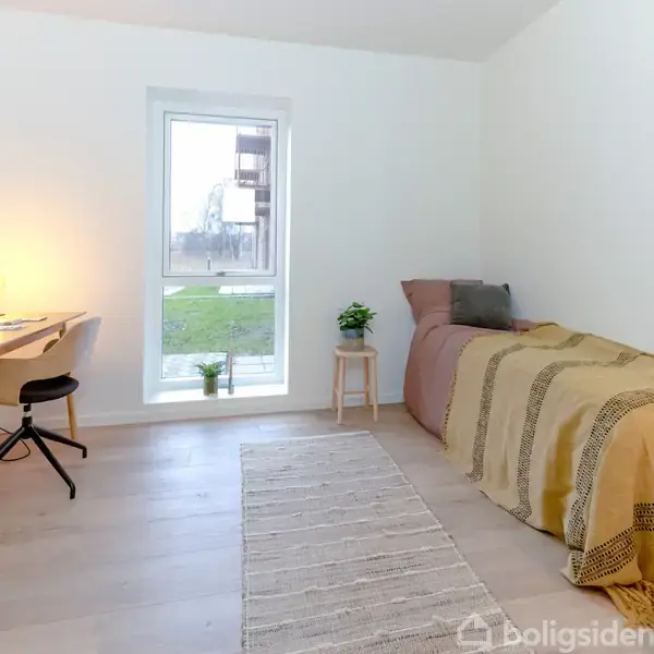 Et enkelt værelse har en seng med tæppe i højre hjørne, et skrivebord med lampe og stol til venstre. Det har en lys væg og et stort vindue.