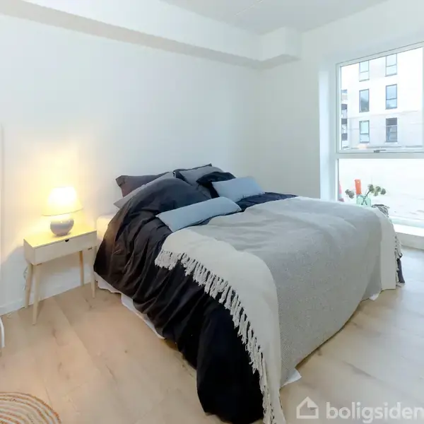 En dobbeltseng med mørk sengetøj og plaid står i et lyst soveværelse. Et natbord med lampe ved siden af, og et vindue med udsigt til en bygning udenfor.