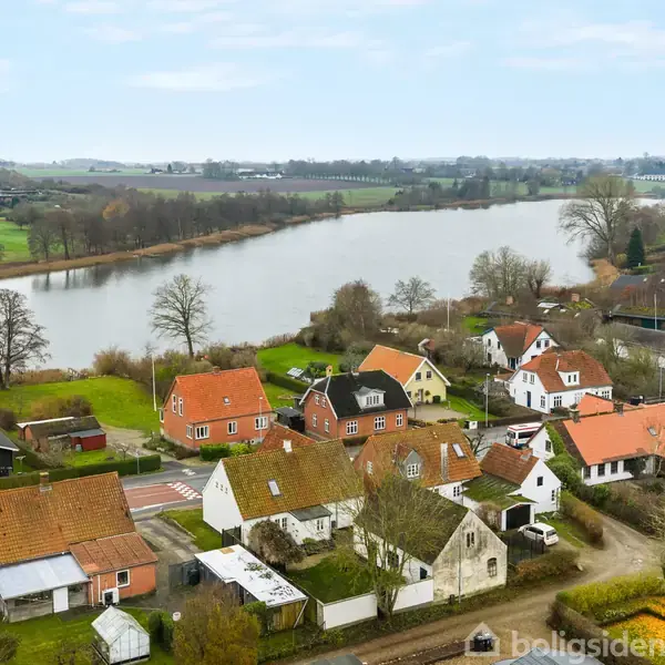 Flere boliger med røde tage ligger tæt ved en snoet flod, omgivet af grønne marker og træer, under en blå himmel.