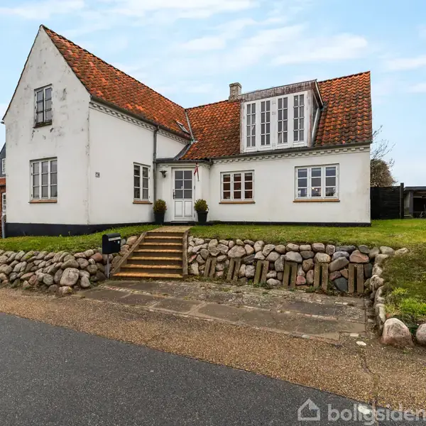 Et hvidt hus med rødt tegltag står på en forhøjning med stentrappe og stenmur omkring. Ved siden af ligger en græsplæne, og i baggrunden ses flere huse.