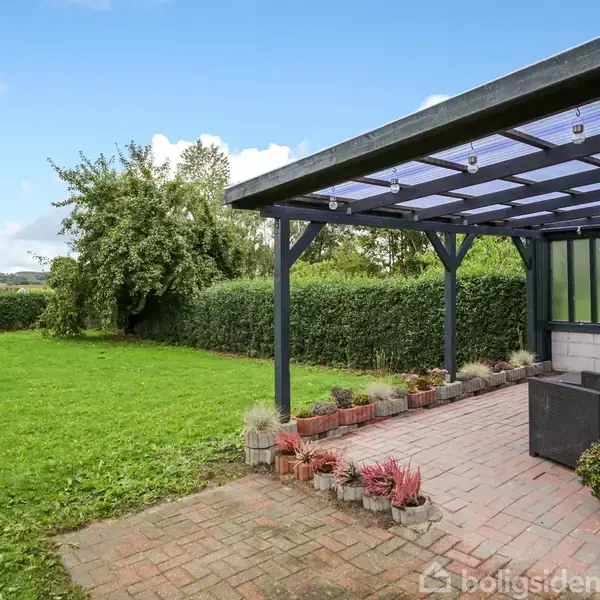 Terrasse med sorte møbler under en halvåben overdækning. Flisebelægningen omringes af planter i krukker. Græsområde og hække omgiver terrassen i en landlig have.