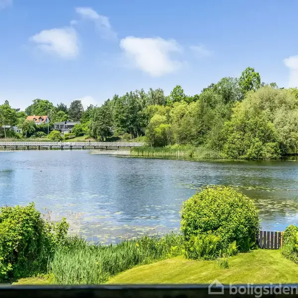Sø omgivet af frodige buske og træer under en blå himmel med få skyer. I baggrunden ses en lille bro og flere huse.