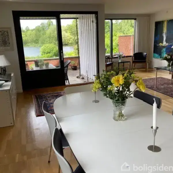 Huset med rødt tegltag står stille i en frodig have med veltrimmede buske ved siden af en sø. Omkranset af grønne områder og et moderne nabohus.