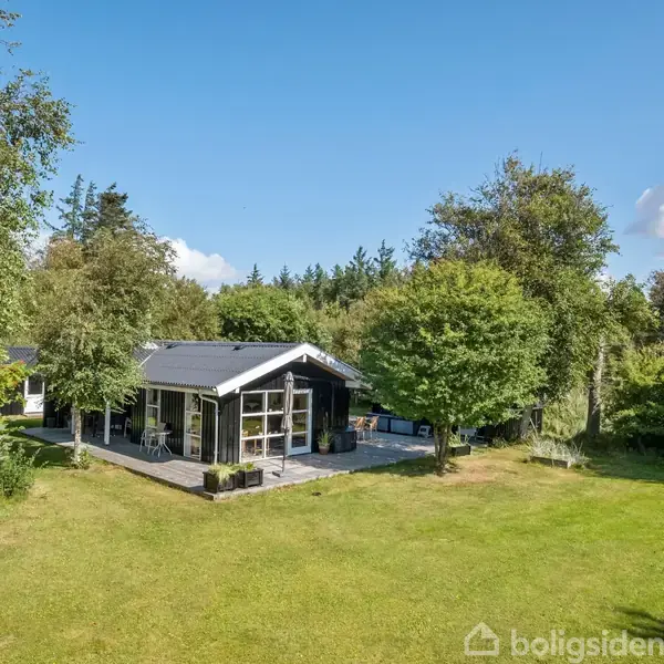 Et træhus står stille omgivet af grønne træer og en græsplæne under en blå himmel. Store vinduer vender ud mod en solbeskinnet terrasse i et stille naturområde.