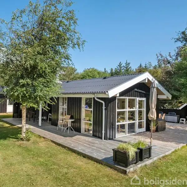 Sort træhus står stille omkranset af frodige træer. Udendørsterrassen prydes af planter, stole og et bord, mens en parasol står ved siden af. Græsplænen omkranser huset.