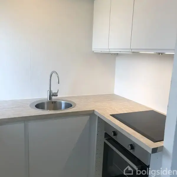 En moderne køkkenvask af rustfrit stål er monteret i en lys træbordplade. Ved siden af er en sort kogeplade placeret. De hvide skabe hænger over bordet.