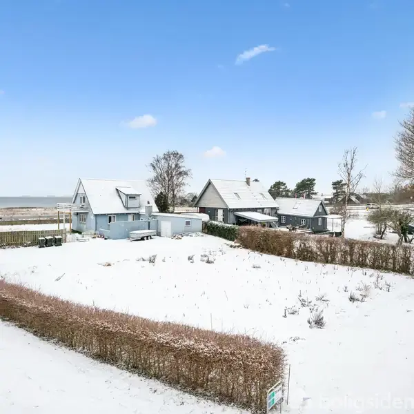 To huse dækket af sne står ved siden af hinanden på en åben grund med hæk, omgivet af træer, under en klar blå himmel.