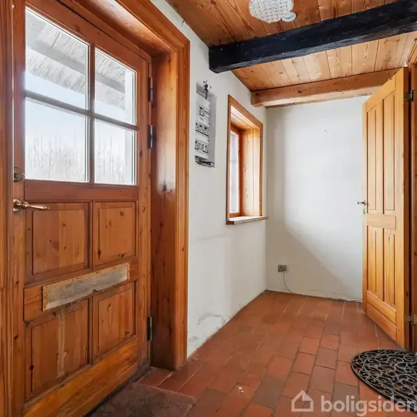 En trædør fører ind i en gang med rødlige fliser. Til venstre er der et vindue og en træbeklædt loft med synlige bjælker. En dørmåtte ligger ved siden af en åben dør.
