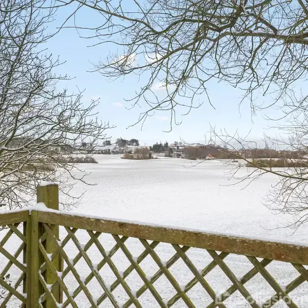 Træhegn står stille, dækket af sne. Baggrunden viser et snedækket landskab med marker og træer, mens himlen er klar og blå. Desuden er der spredte huse i det fjerne.