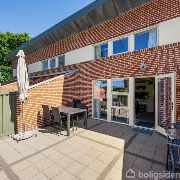 Murstenshus med åbne terrasse-døre; sorte udemøbler står på flisebelagt terrasse. Grønne træer i baggrunden. Et skilt ved døren viser tallet "3".
