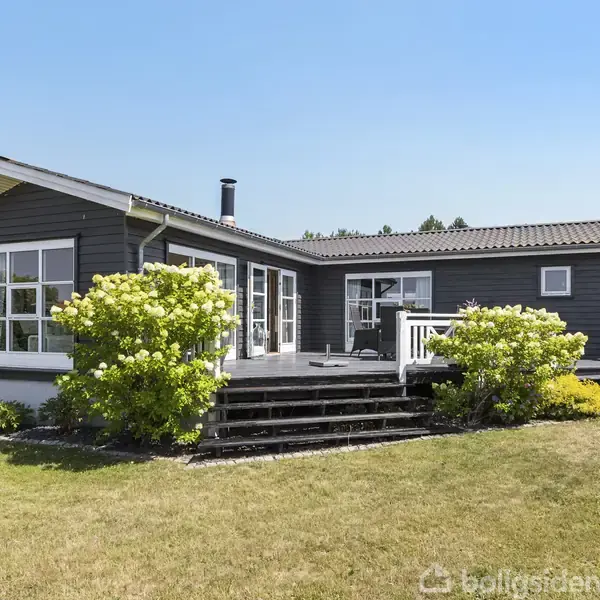 Et moderne, mørkegråt hus med store vinduer står stille i en velplejet have. En terrasse med trapper fører op til hoveddøren, omgivet af grønne buske i solrigt vejr.