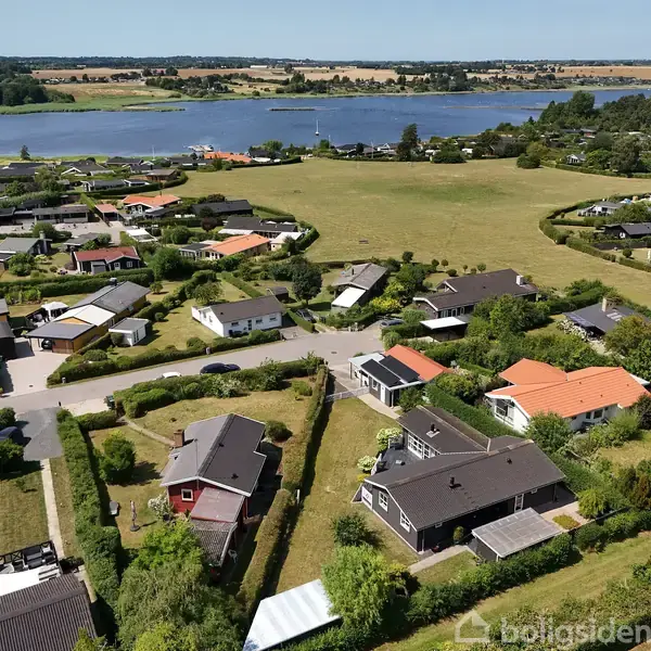 En række huse med haver ligger langs veje. De befinder sig i et åbent område omgivet af marker, med en stor sø i baggrunden under en klar blå himmel.