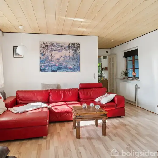 Rød sofa står i et lyst stueområde med træloft. Sofabordet har glas og dekorative genstande. Et stort maleri hænger på væggen, omgivet af grønne planter ved vinduet.