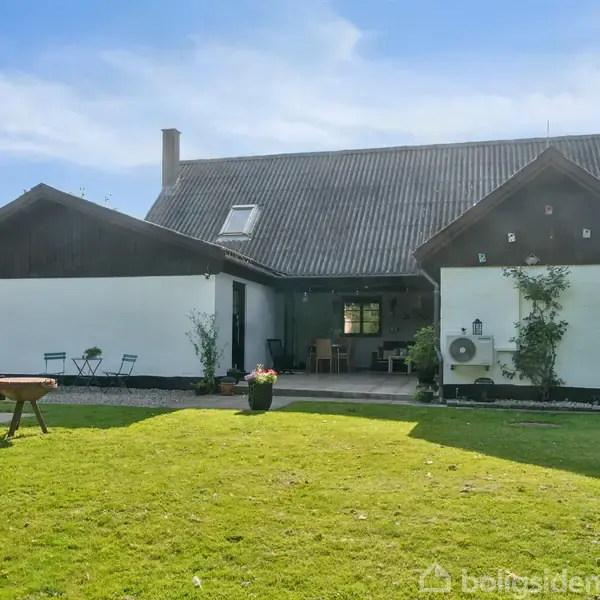 En hvidkalket villa med mørkt tag står i en grøn have. Terrassemøbler og planter er placeret foran huset. Himlen er blå med spredte skyer.