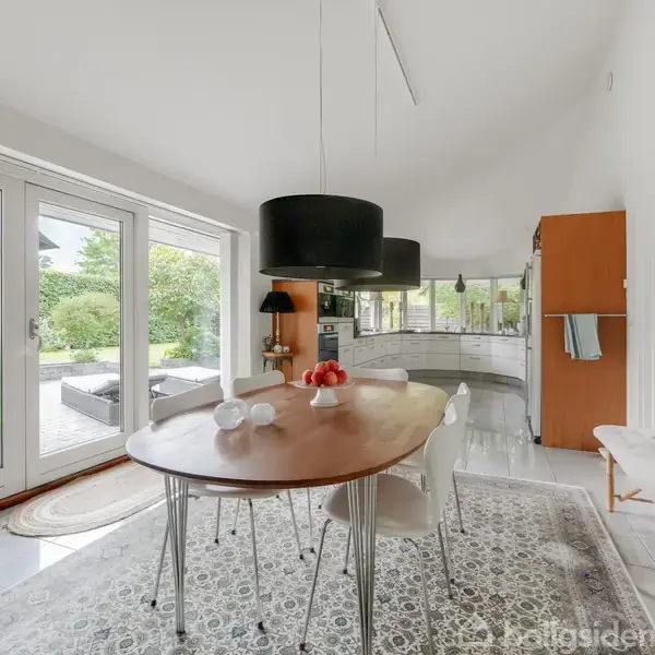 Et ovalt spisebord står i en lys spisestue med store glasdøre til en terrasse. Moderne lamper hænger over bordet. Køkkenet er synligt i baggrunden med hvide skabe.