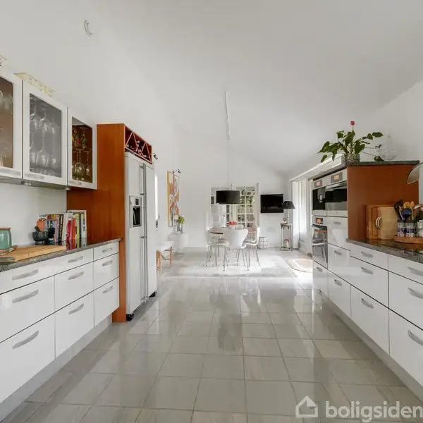 Et lyst, moderne køkken med hvidt interiør har skabe fyldt med glas. Langs væggene står køkkenskabe og apparater. I baggrunden ses et spisebord i et rummeligt, åbent område.