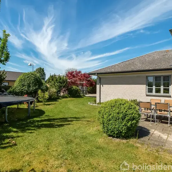 En villa omkranset af frodig have med græsplæne og træer. Terrasse med bord og stole ved siden af huset. Trampolin placeret i haven under en blå, skyet himmel.