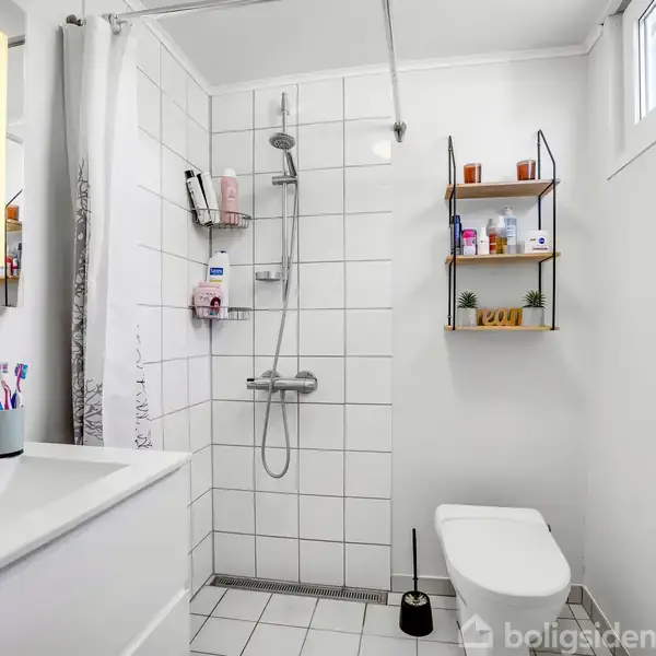 Badeværelse med brusekabine og hvidt flisegulv. Håndvask med tandbørster til venstre. Toilettet står til højre under et væghængt hyldesystem. Tekst: "GOOD VIBES ONLY," "BELIEVE," "DREAM," "CREATE," "IMAGINE," "LOVE," "TAK."