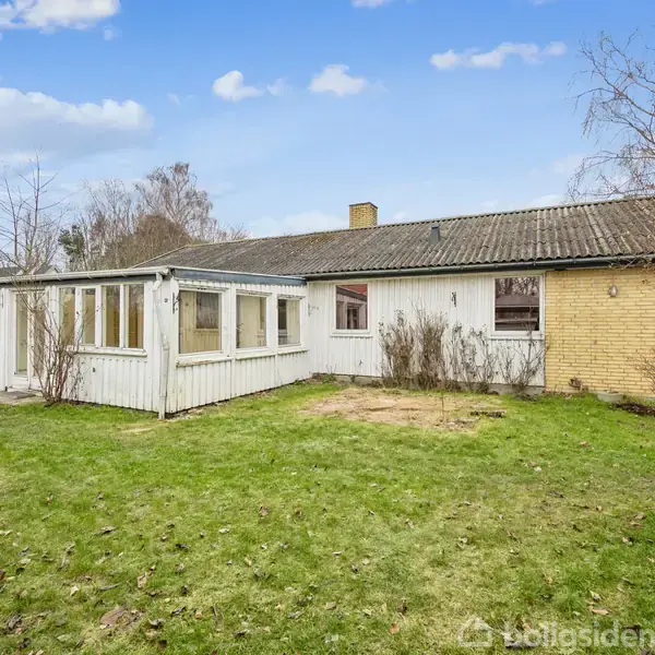 Et lavt, langstrakt hus med hvide træpaneler og et sort tegltag står i en grøn have med spredte buske og træer under en blå himmel.