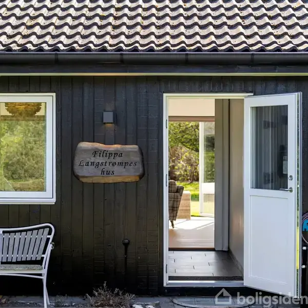 En sortmalet husfacade med et skilt, hvor der står "Filippa Langstrømpes hus". En åben dør fører til en lys stue med udsigt til en grøn have.
