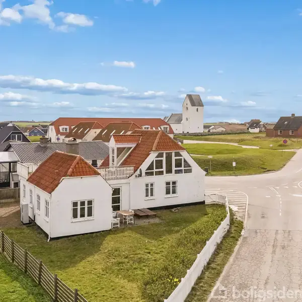 Et hvidt hus med rødt tag står stille i et åbent, naturskønt område nær en vej og andre huse. Himlen er blå med hvide skyer.