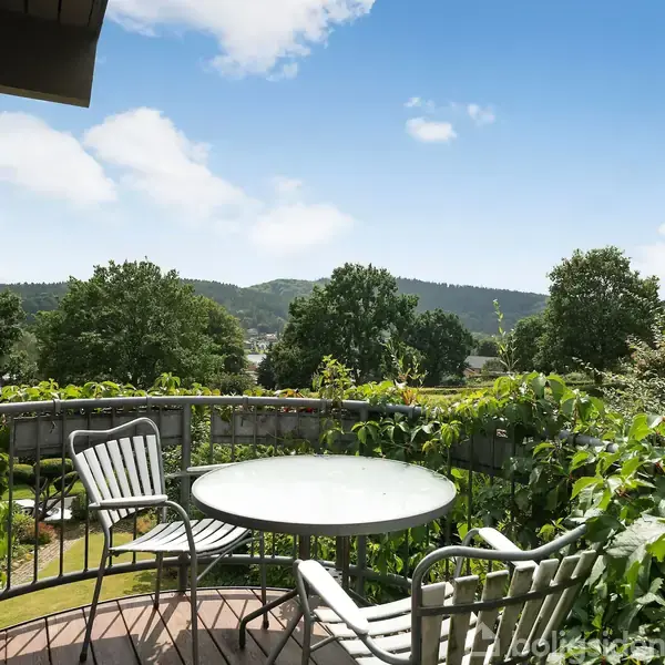Rundt bord og tre stole står på en altan omkranset af grønne planter. I baggrunden ses en frodig have og skovklædte bakker under en blå himmel med få skyer.