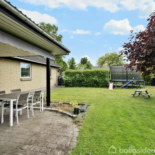 Terrassebord med stole står på en flisebelagt terrasse ved et hus. I haven ses et træ, en trampolin og en rutsjebane. Baggrunden har grønne buske og en blå himmel.