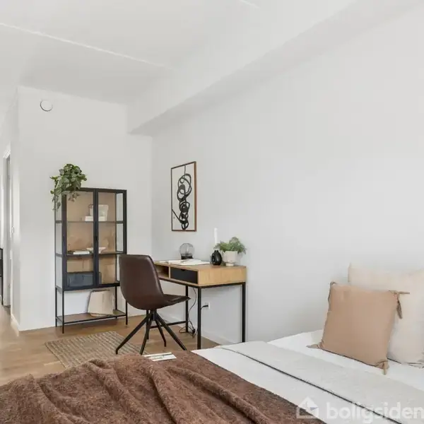 Et lyst soveværelse med en seng dækket af hvide og brune puder, står ved siden af et skrivebord og stol. En åben reol med planter og kunst pryder væggen.