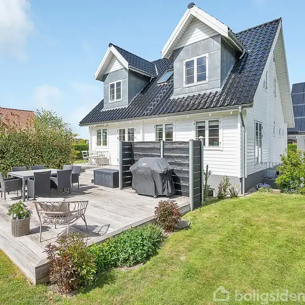 Hvidt hus med sorte tagplader, omgivet af velplejet græsplæne. Træterrasse med havesæt og grill i forgrunden. Solrigt vejr med få skyer.