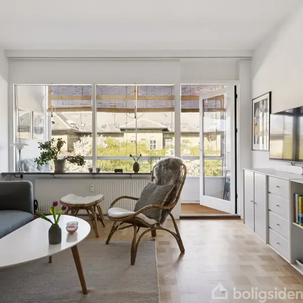 Stue med blå sofa og rundt sofabord, stol med fletdesign nær vindue. Fjernsyn på væggen over reol, der indeholder bøger. Store vinduer og åben altandør viser udsigt til have.