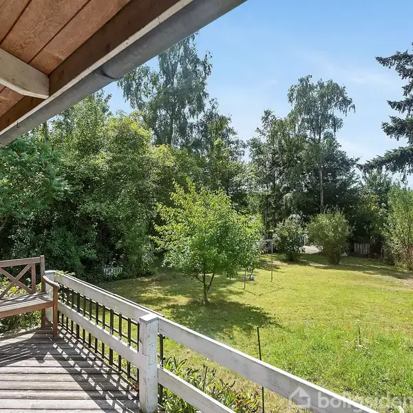 Bænk står stille på en terrasse omgivet af et grønt græsareal og frodig beplantning i en have med klare blå himmel. Ingen tekst er til stede.