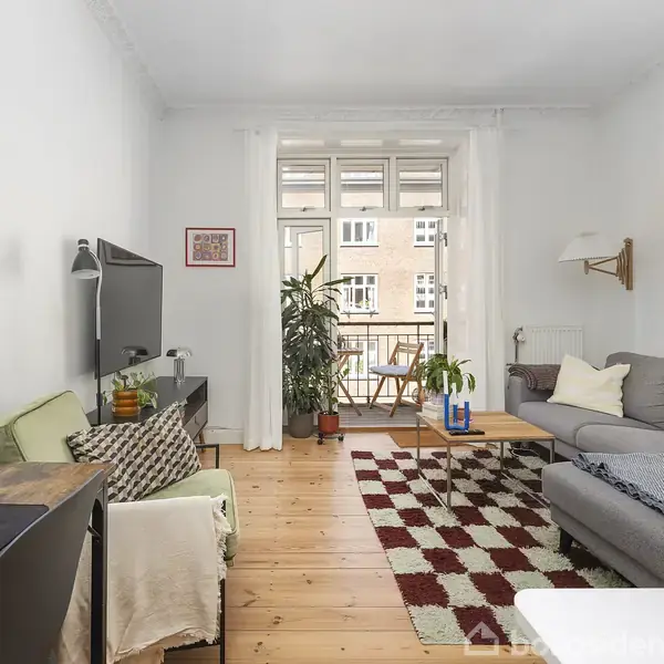 Stue med grå sofa til højre, over et ternet tæppe, og fjernsyn til venstre. I baggrunden er store vinduer med planter. Væggene har kunstværker. Trægulve og lyst interiør.