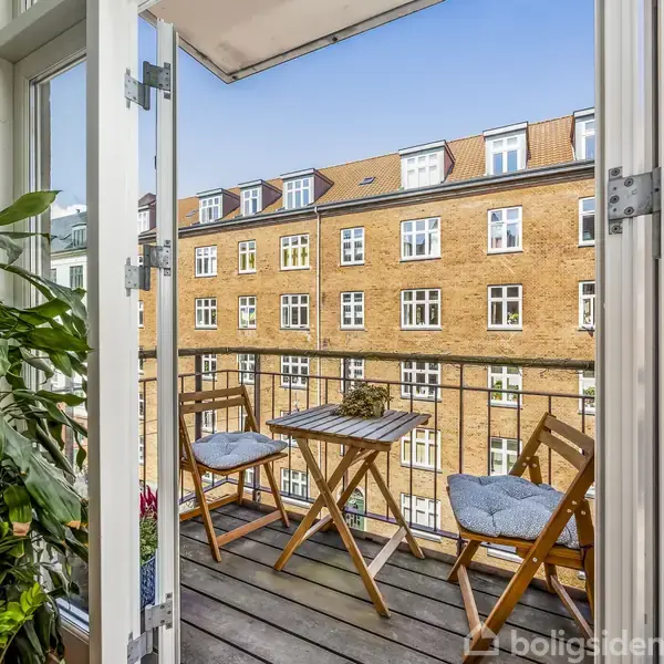 Balkon med to foldestole og et lille bord, omgivet af en plante. Udsigt over en rødstensejendom med flere vinduer under blå himmel.